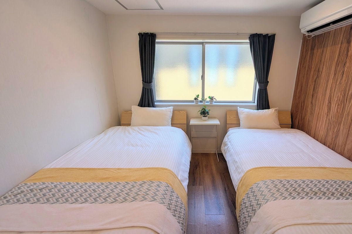 A hotel room with two beds, a small bedside table with a lamp, a window with curtains, and an air conditioning unit.