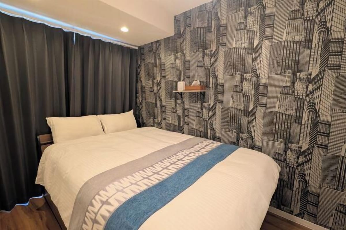A hotel room with a large bed, dark curtains, a cityscape wallpaper, and a small shelf with items.