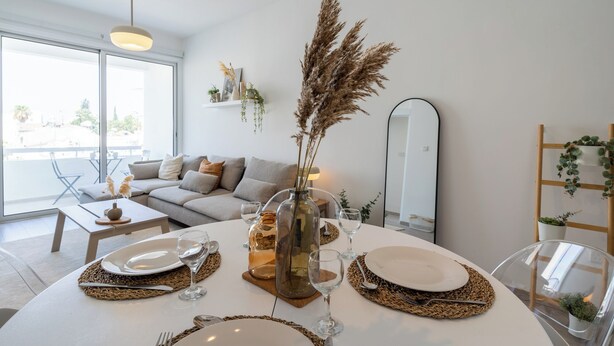 A modern living room with a dining area set for a meal, featuring a sofa, a dining table, and a large window. - Hermes 2-BR Apt. & Balcony in Larnaca (Larnaca)