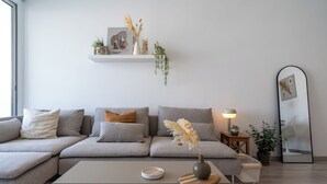 A modern living room with a sectional sofa, a coffee table, a side table with a lamp, a mirror, and a wall-mounted shelf with decorative items. - Hermes 2-BR Apt. & Balcony in Larnaca (Larnaca)