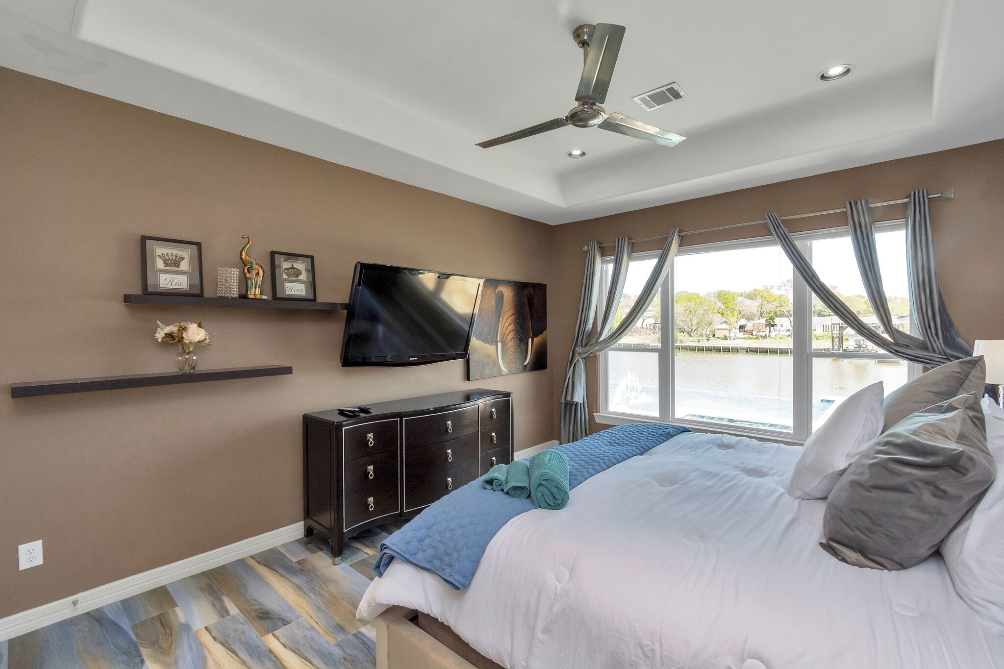A bedroom with a bed, a TV, a ceiling fan, a dresser, and a window with curtains.