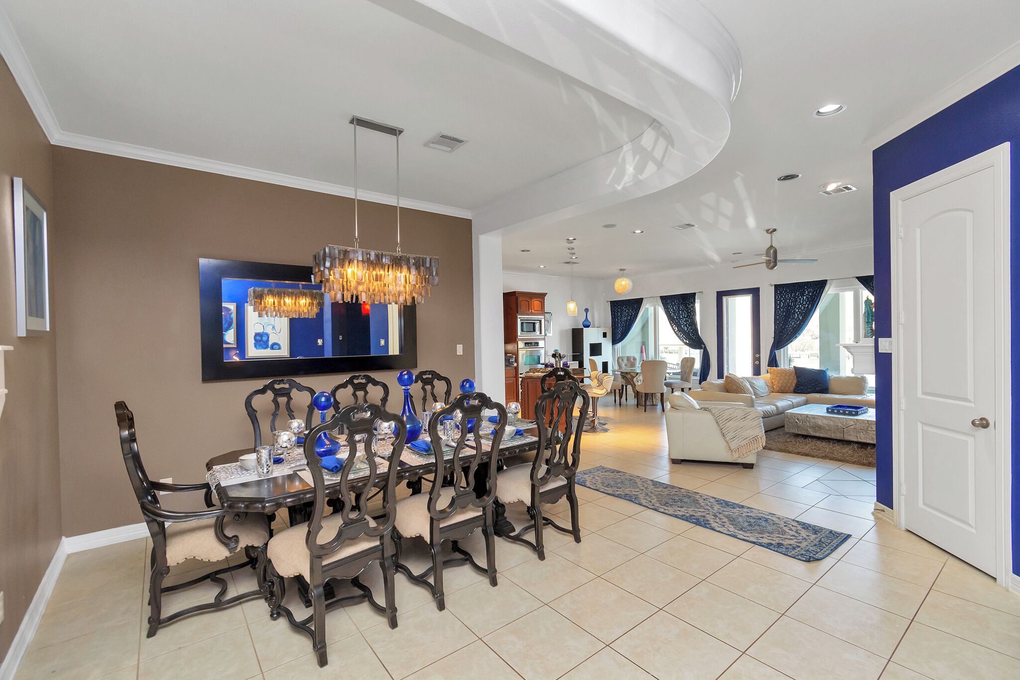 A spacious dining area with a modern chandelier, a large flat-screen TV mounted on the wall, and a view into the adjacent living room.