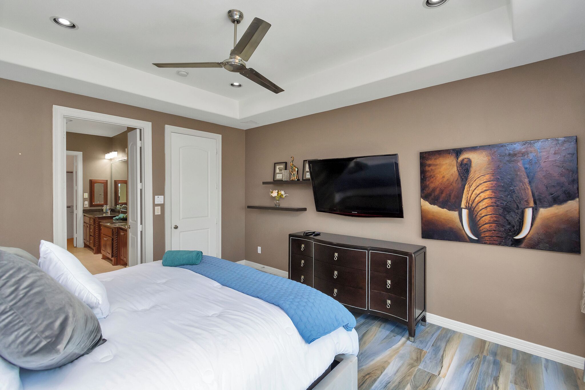 Un dormitorio con cama, ventilador de techo, televisor, cómoda y obras de arte en la pared.