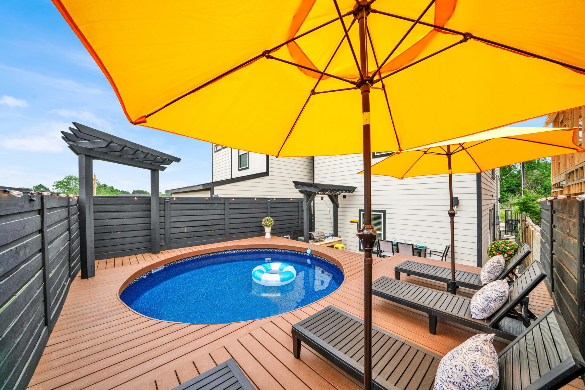 A backyard with a swimming pool, wooden deck, outdoor furniture, and a large yellow umbrella.