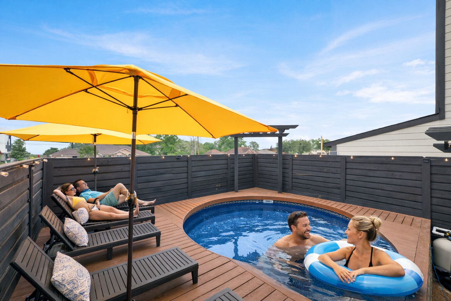 En baghave med swimmingpool, træterrasse, liggestole og en gul parasol.
