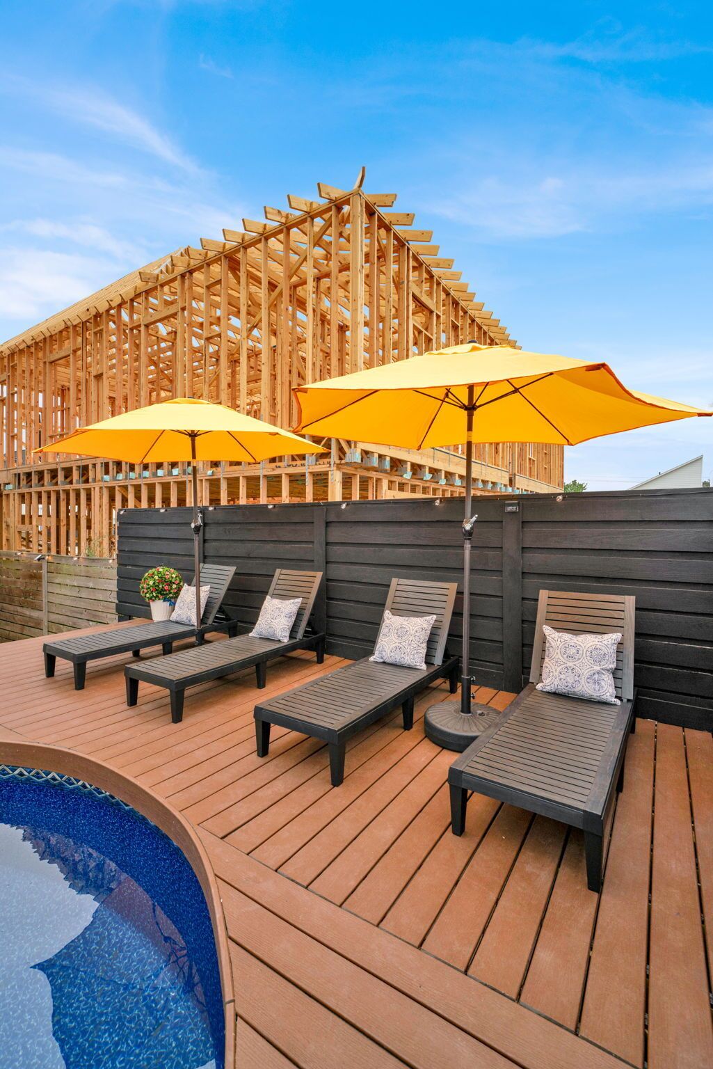 A wooden deck with lounge chairs and yellow umbrellas, a swimming pool, and a wooden pergola structure in the background.