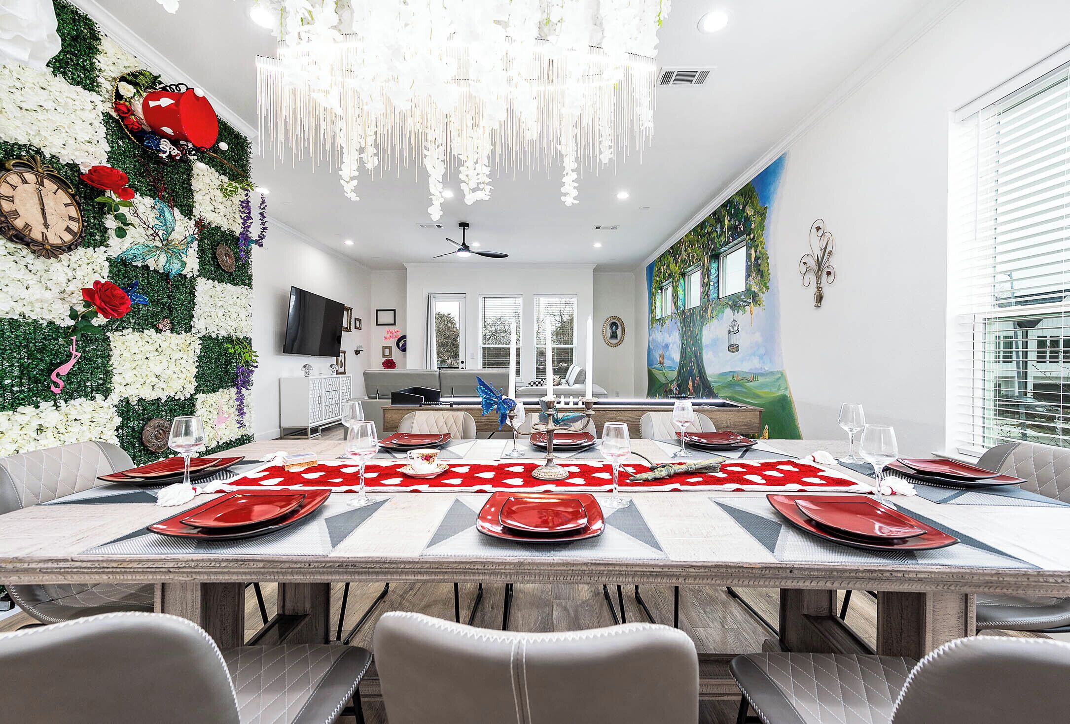 A dining area with a long table set for a meal, a kitchen in the background, and a mural on the wall.