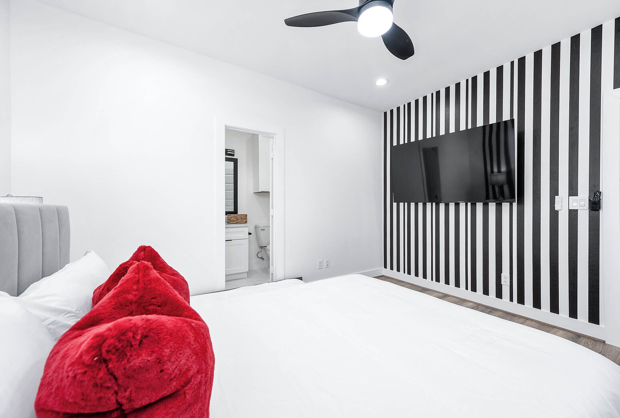 Quarto moderno com cama, ventilador de teto, TV presa a parede listrada e banheiro visível através de uma porta aberta.