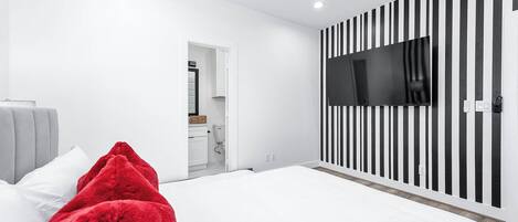 Quarto moderno com cama, ventilador de teto, TV presa a parede listrada e banheiro visível através de uma porta aberta.