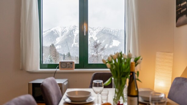A dining table with plates, glasses, and a bottle, set against a window with a view of snow-capped mountains. - Centrally located and at the ski slope. (Hohentauern)