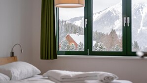 A bedroom with a bed, a window with a view of a snowy mountain and a cabin, and a green curtain. - Centrally located and at the ski slope. (Hohentauern)