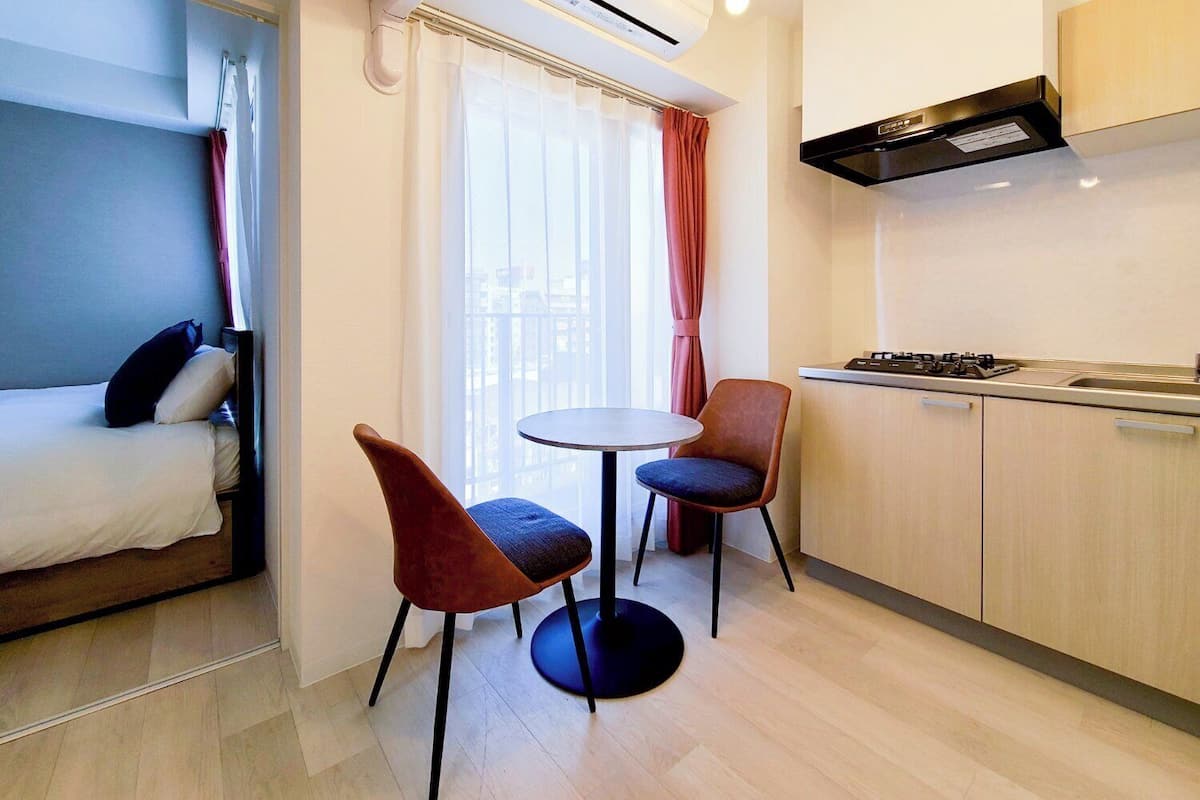 A compact hotel room with a bed, a small table, two chairs, a kitchen area with a stove, and a window with curtains.