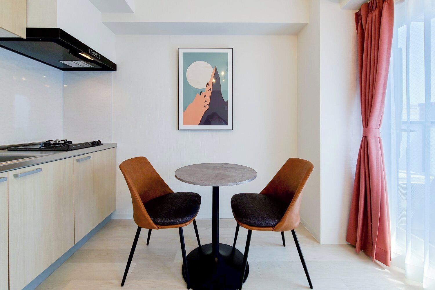 A modern kitchen with a gas stove, two chairs, a small round table, and a framed artwork on the wall.