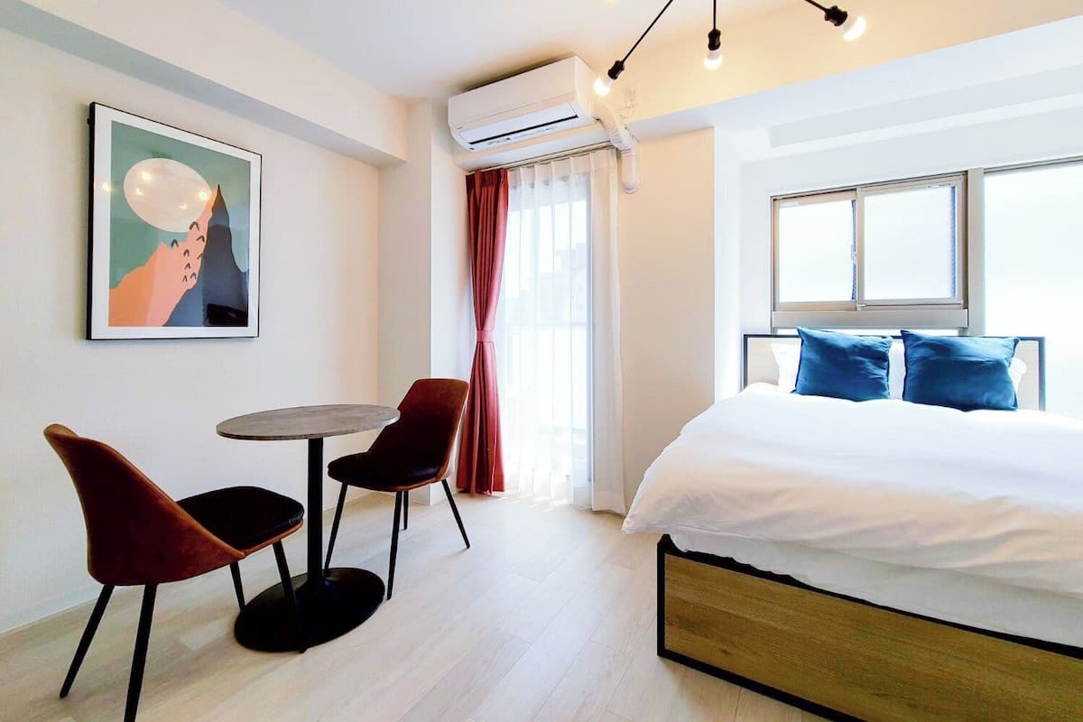 A hotel room with a bed, two chairs, a small table, a painting, an air conditioning unit, and a window with curtains.