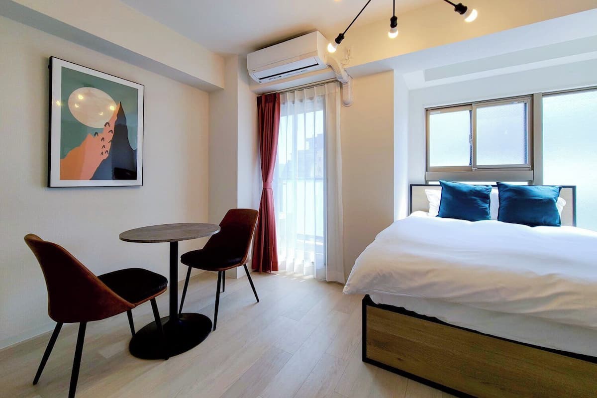A hotel room with a bed, a small table with two chairs, an air conditioning unit, a window with curtains, and a framed picture on the wall.