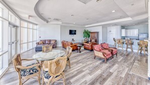 A spacious living area with a round glass table, wicker chairs, leather sofas, and a kitchen in the background. - Legacy Tower II- #1406 - 2BR/2BA Penthouse Gulf-View Condo - Resort Pools + Spa (Gulfport)