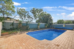 A swimming pool with a brick patio, surrounded by a fence and greenery.