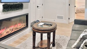 Ein modernes Wohnzimmer mit Flachbildfernseher, Kamin, rundem Holztisch und grauem Sofa.