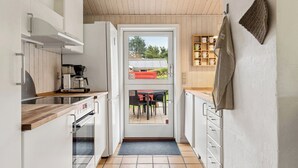 Eine schmale Küche mit Holzbalkendecke, einer Kaffeemaschine und Blick durch eine Glastür auf einen Außenbereich mit Sitzgelegenheiten.
