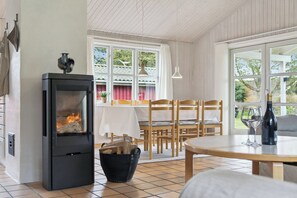 A cozy room with a wood-burning stove, a dining table, chairs, and large windows. - Fijne vakantiebasis bij strand en Spodsbjerg (Rudkøbing)