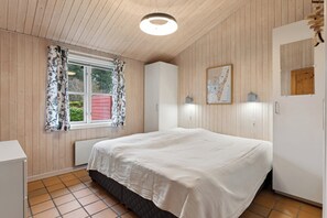 A bedroom with wooden panel walls, a bed with a white bedspread, a window with floral curtains, a map on the wall, and a radiator. - Fijne vakantiebasis bij strand en Spodsbjerg (Rudkøbing)