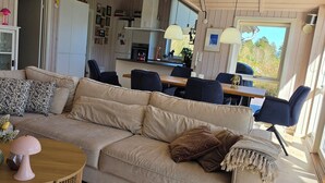 A living room with a sofa, a dining table, chairs, a kitchen in the background, and a window with a view.