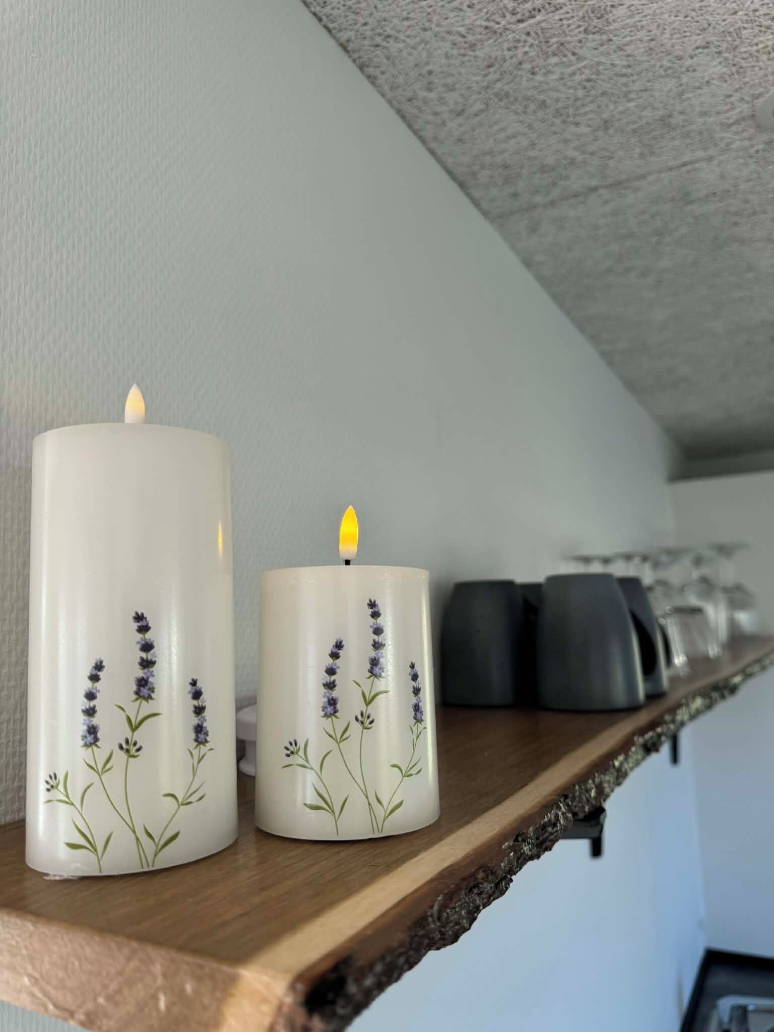 Zwei weiße Kerzen mit Lavendel-Motiven auf einem Holzregal.