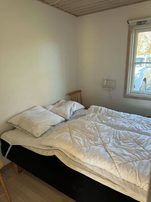 Un lit simple avec une literie blanche, une tête de lit en bois, une fenêtre donnant sur une voiture et une prise de courant fixée au mur.