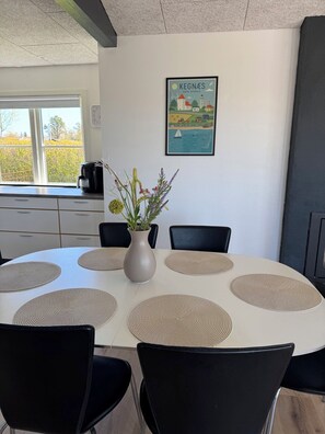 A round table with six chairs, a vase with flowers, a framed picture of Kegnæs, and a window with a view of greenery. - Lovely holiday home with beautiful views (Sydals)