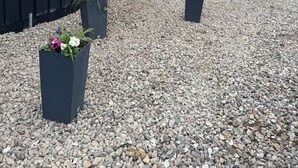 Rakennuksessa on musta julkisivu, valkoiset koristeet ja kolme ikkunaa. Sen ympärillä on sorapolkua ja kukilla koristeltuja ruukkukasveja.