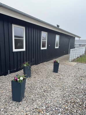 A building with a black exterior, white trim, and three windows. There are gravel paths and potted plants with flowers. - Lovely holiday home with beautiful views (Sydals)