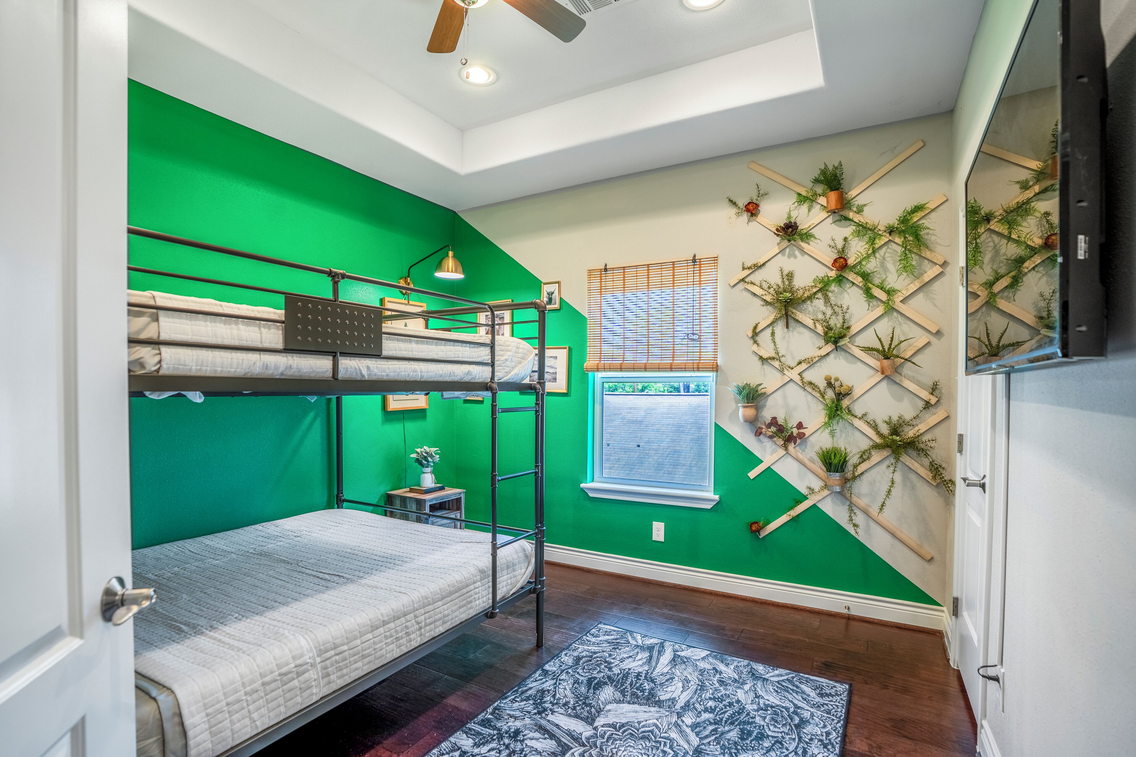 A room with bunk beds, a small table with a lamp, a window with blinds, and a wall-mounted plant display.