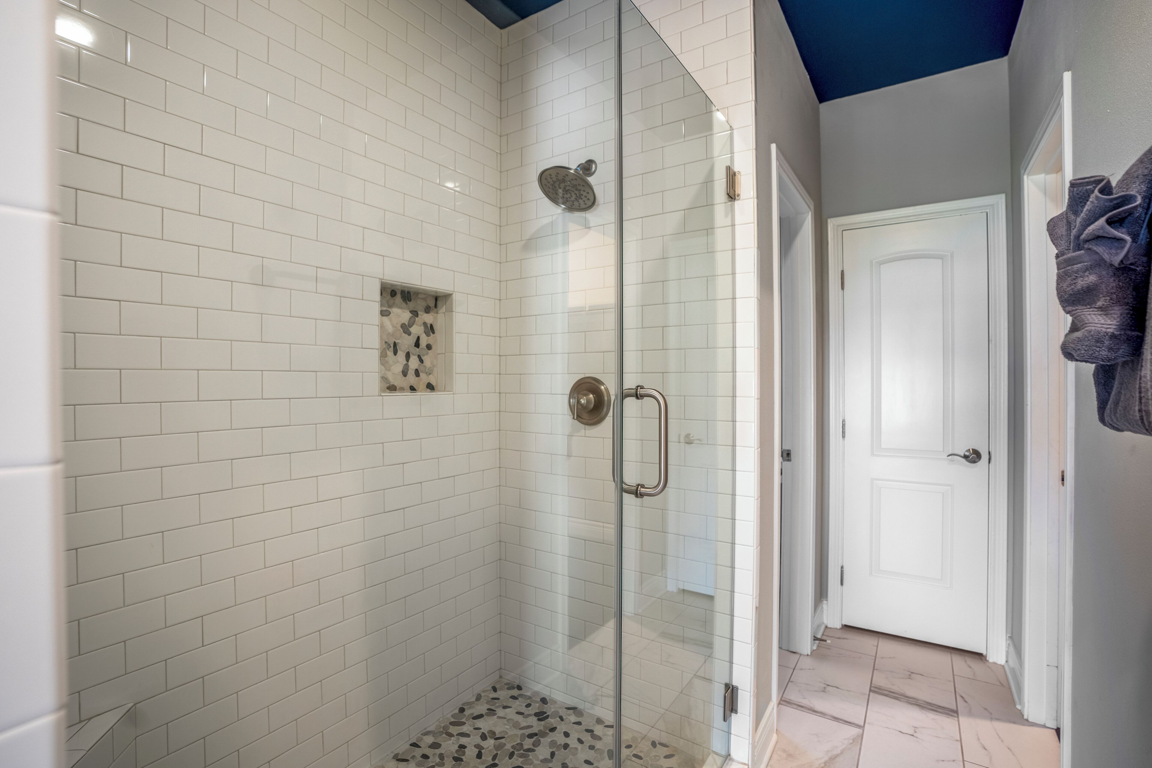 A glass-enclosed shower with white subway tiles, a small decorative tile accent, and a door leading to a hallway with a white door and a towel rack.