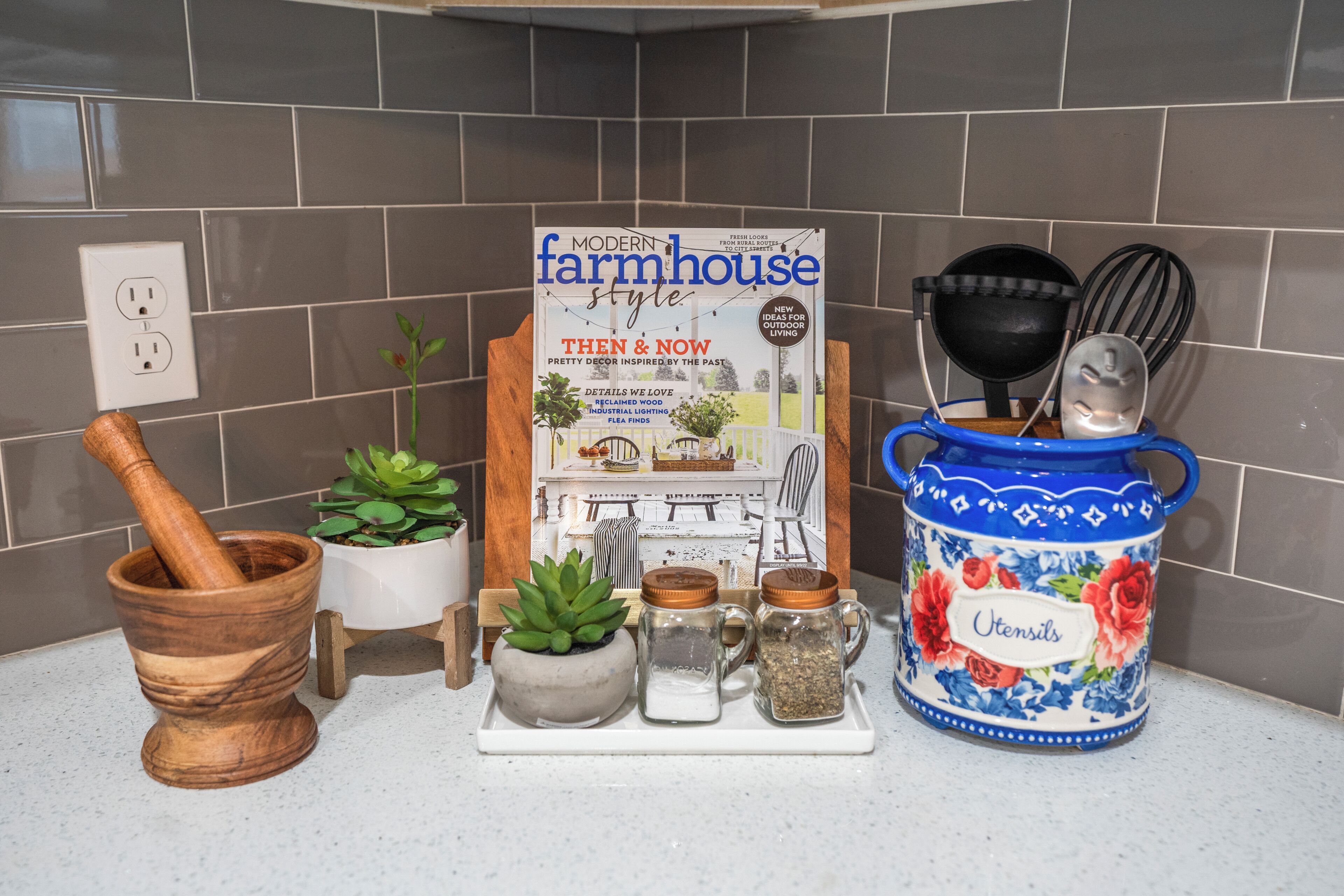Una encimera de cocina con una revista, un mortero y una maja, una planta en maceta y un tarro de cerámica que dice 'Utensils'.