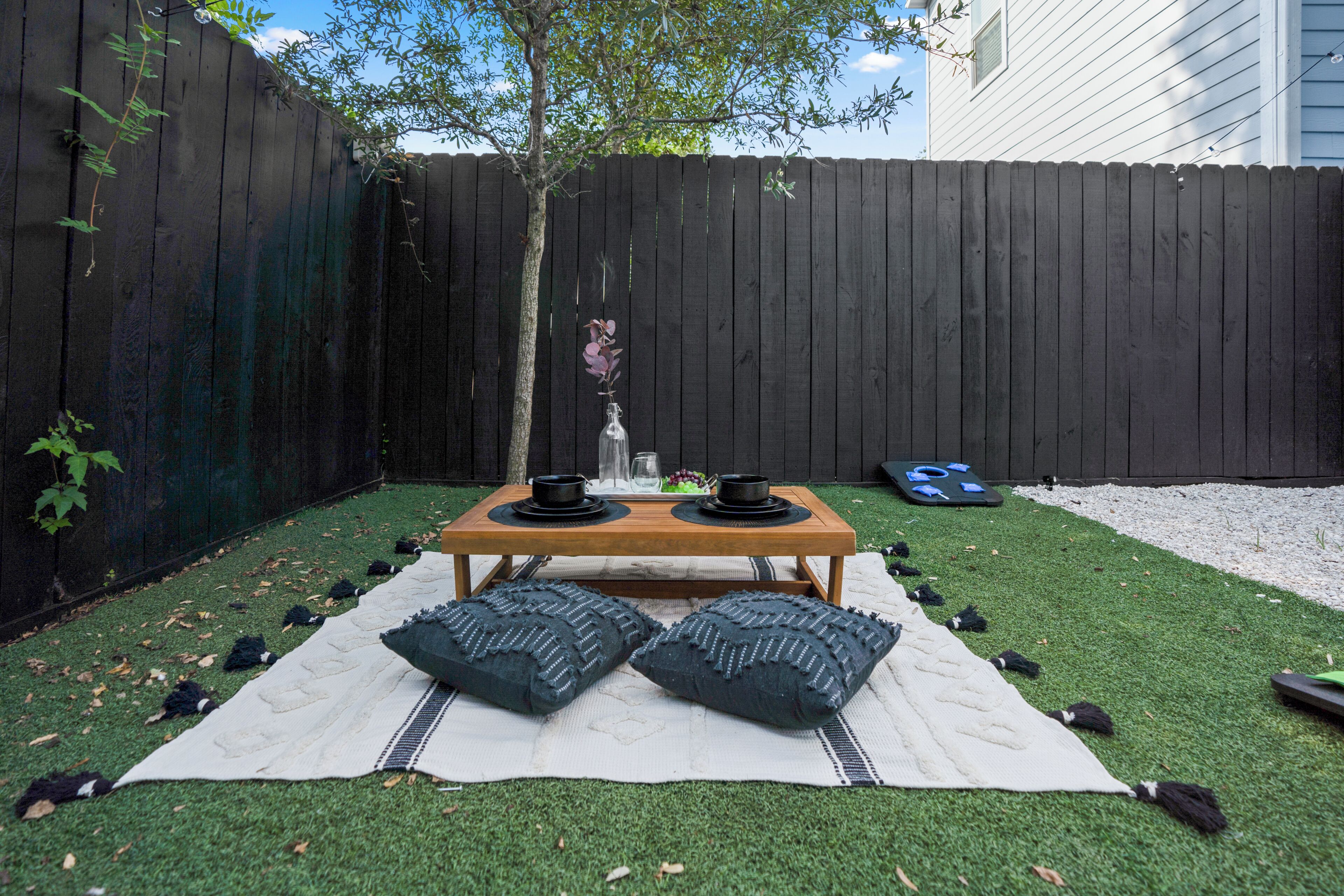 A backyard with a wooden table set for two, surrounded by a white picnic blanket and two decorative pillows.