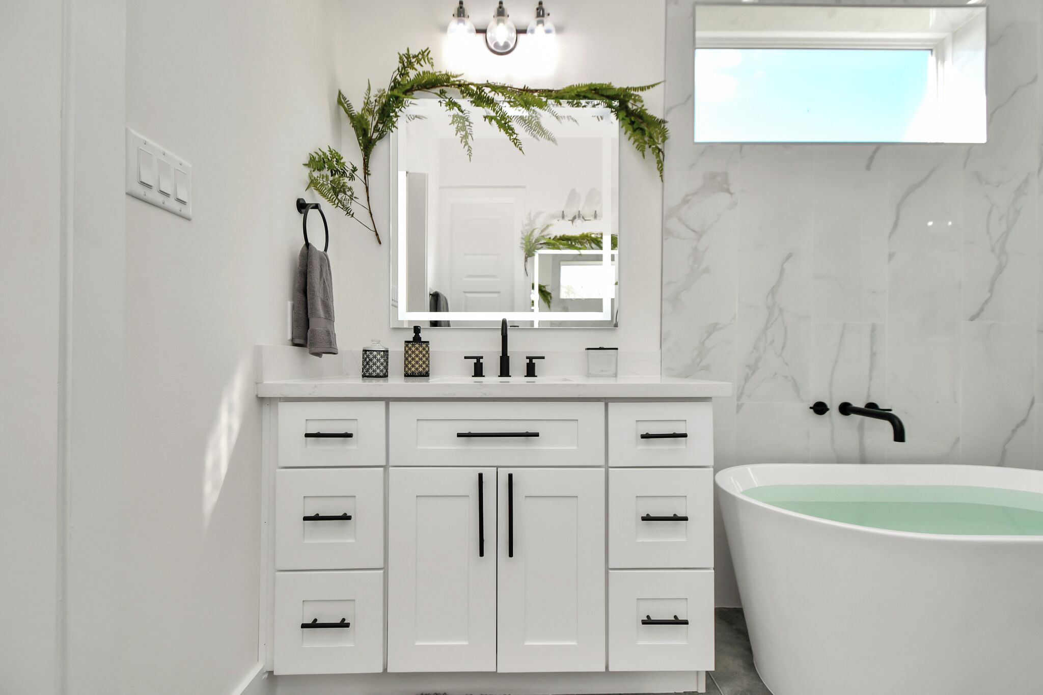 A modern bathroom with a white vanity, a large mirror, a bathtub, and a light fixture above the mirror.