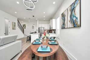 A modern kitchen with white cabinets, a stainless steel refrigerator, and a dining area with a wooden table and blue placemats. - Empress Stylish Retreat (Houston)