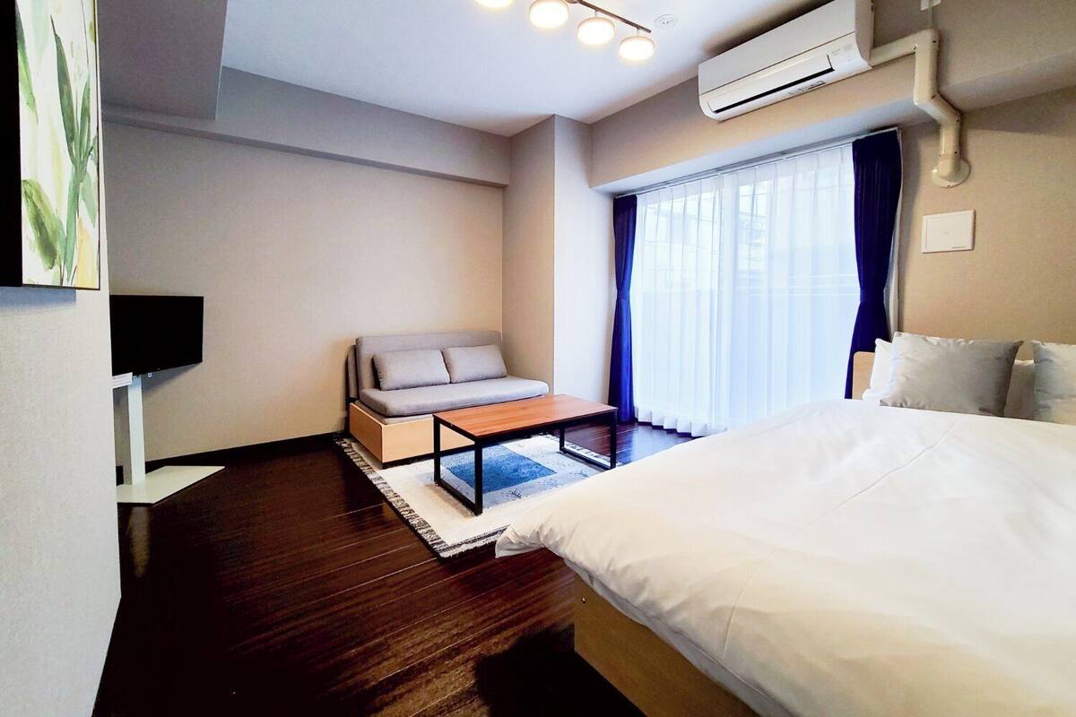 A hotel room with a bed, a sofa, a small table, a TV, and an air conditioning unit.