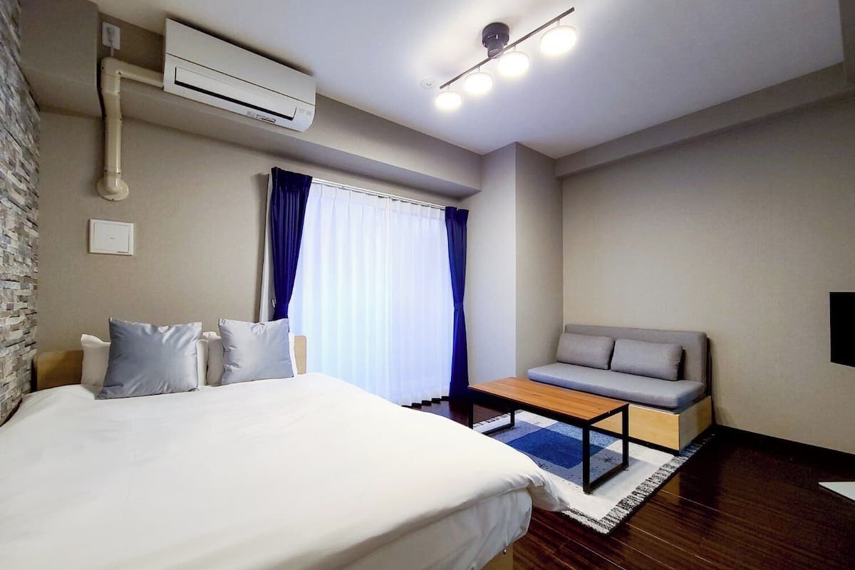 A hotel room with a bed, a sofa, a small table, an air conditioning unit, and a window with curtains.