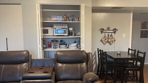 Een woonkamer met een leren bank, een eettafel met stoelen, een keuken met apparatuur en een plafondlamp.