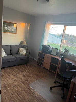 A room with a grey sofa, a desk with a computer, a lamp, and a window with a view of a building and trees.