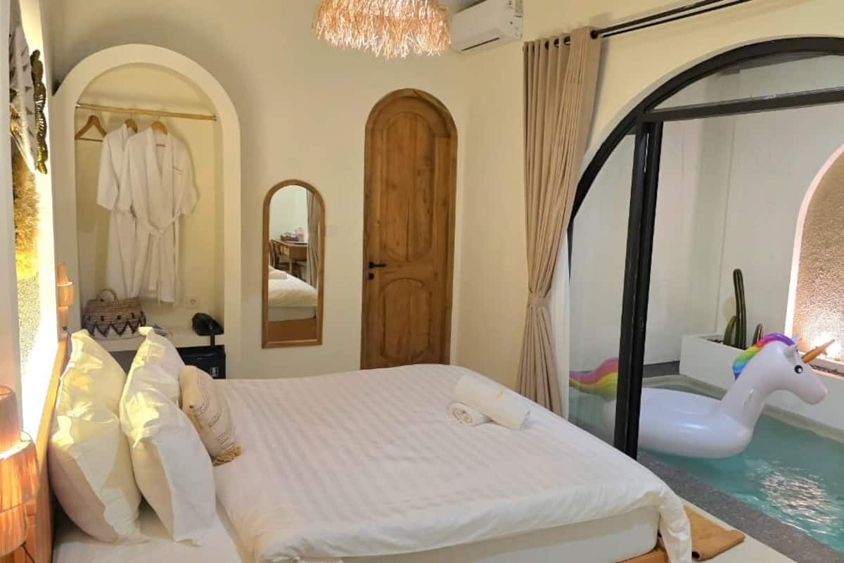 Habitación de hotel con cama, espejo, puerta de madera y vista a una piscina con un flotador con forma de unicornio.