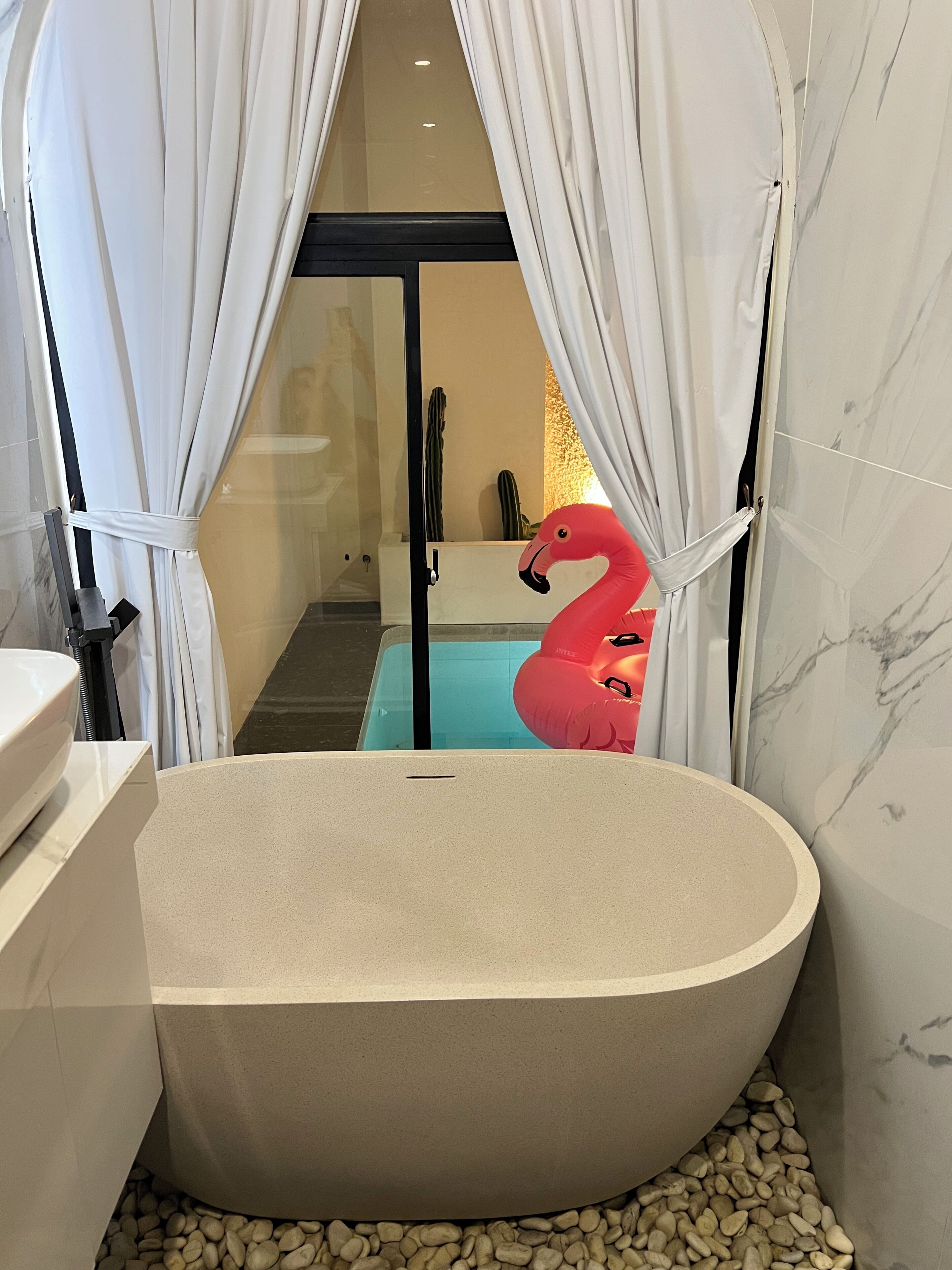 Un bagno moderno con una vasca da bagno indipendente, tende bianche e una decorazione a fenicottero rosso.