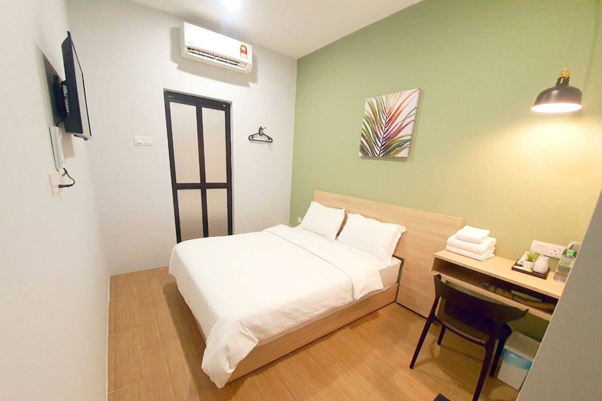 A hotel room with a bed, desk, chair, and a wall-mounted air conditioner.