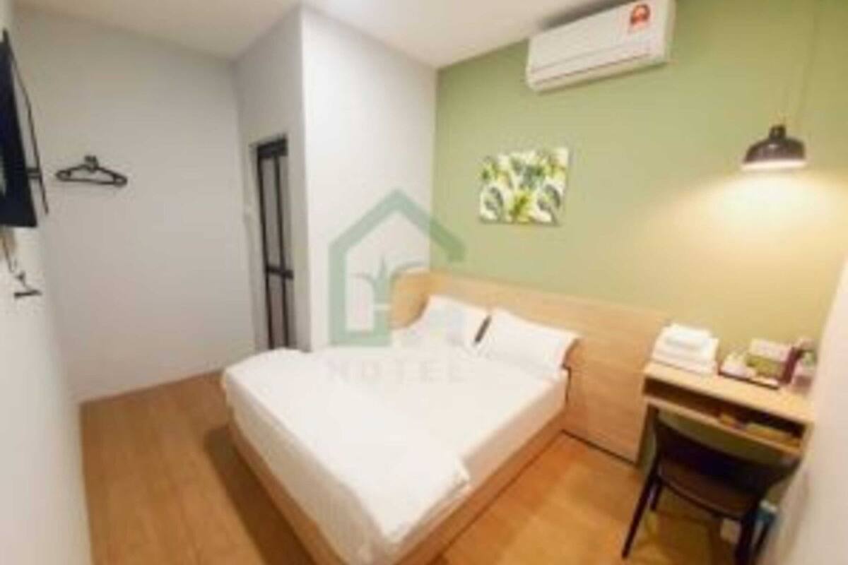 A hotel room with a bed, a desk, and a chair. There is a wall-mounted air conditioner and a small shelf with items on it.