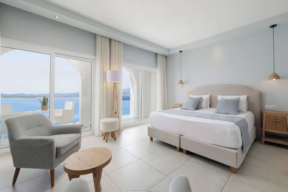 A bedroom with a large bed, a chair, a small table, and a view of the sea.