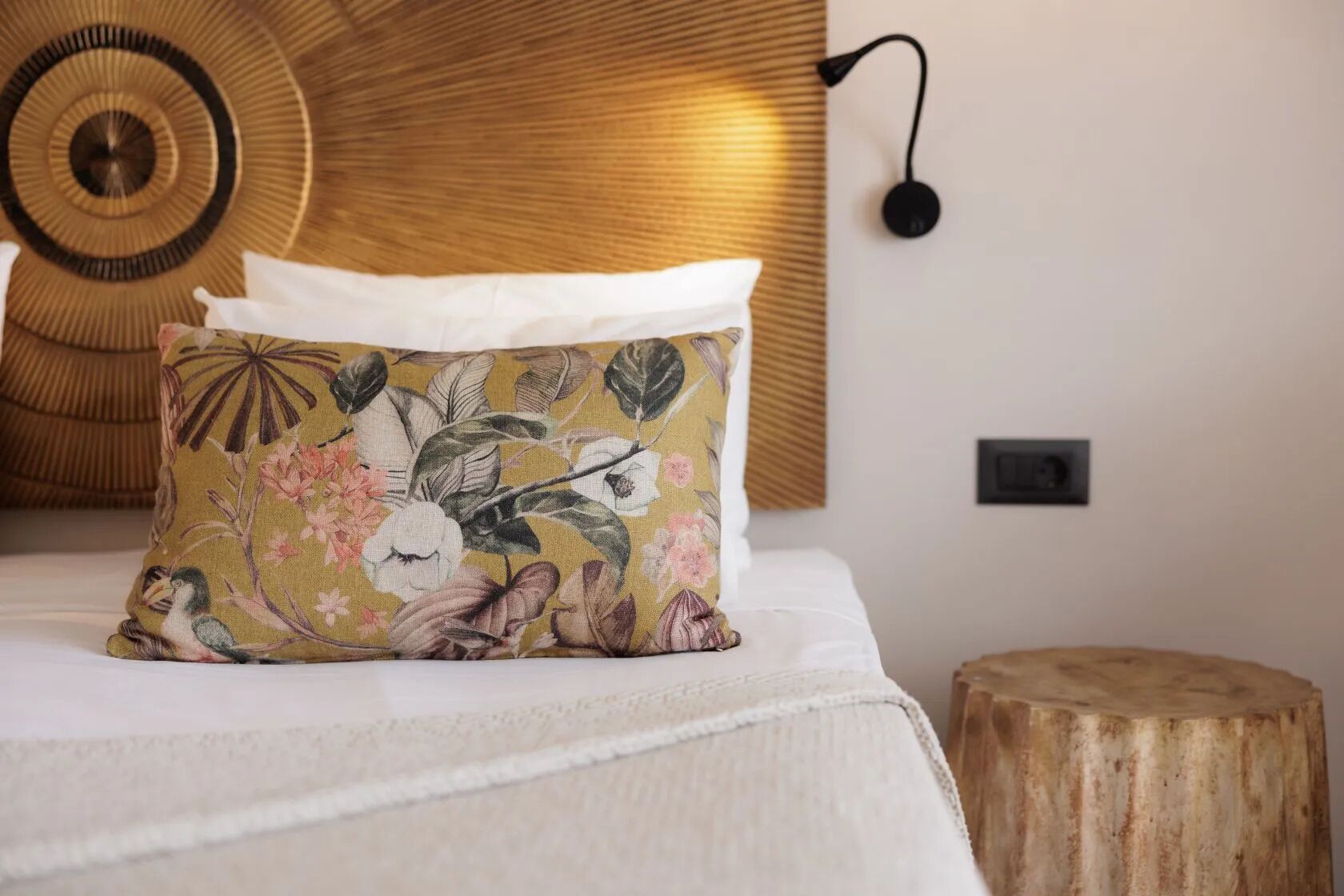 Una cama con un cojín floral, una decoración circular en la pared y un taburete de madera.