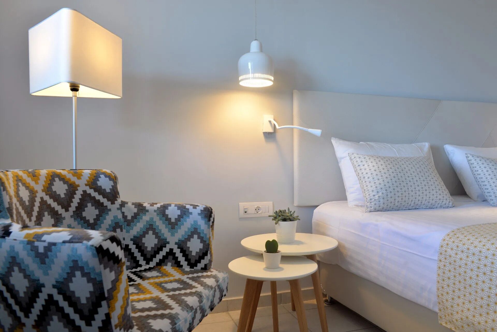 A modern room with a patterned armchair, a small round table with a plant, a bed with patterned pillows, and a wall-mounted lamp.