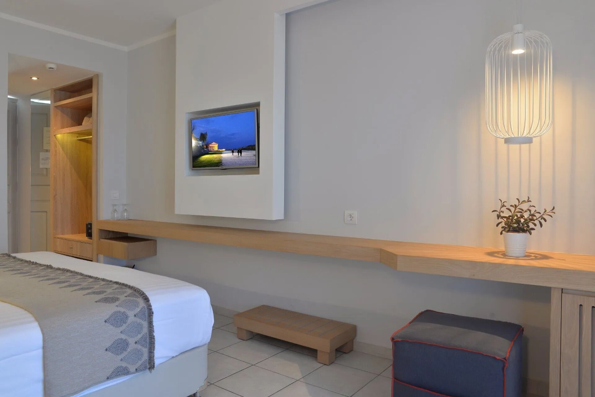A modern hotel room with a bed, a bench, a small table with a plant, a mounted TV, and a pendant light.