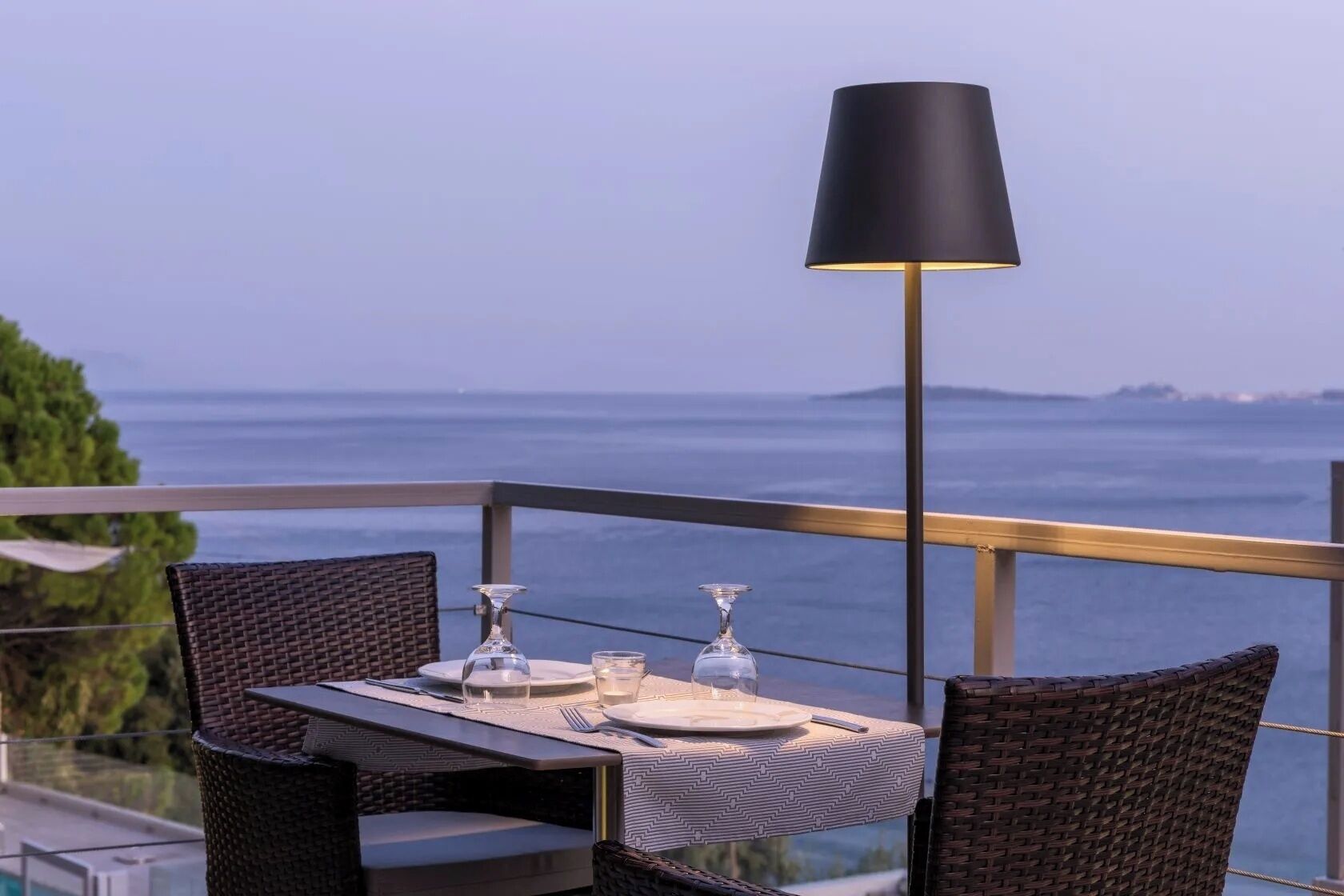 A table set for two with a view of the sea, a black lamp, and wicker chairs.